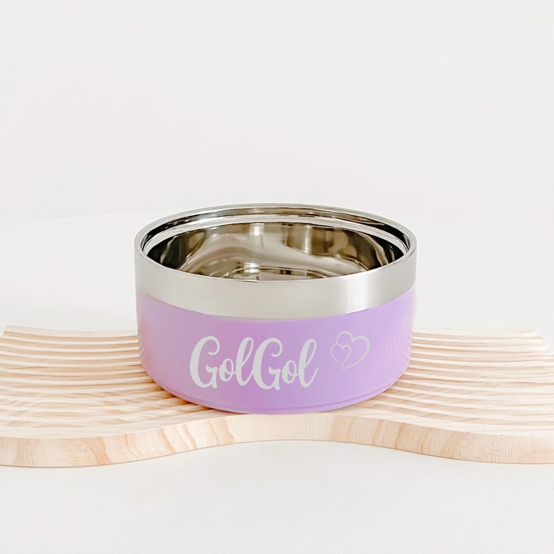 May include: A lavender pet bowl with a stainless steel interior and rim. The bowl has the word "GolGol" and two hearts printed on the side. It sits on a light-colored wooden pet bowl holder.