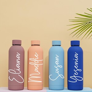 May include: Four personalized water bottles with names "Eliana", "Maclovie", "Susan", and "Yesenia" written in white on the bottles. The bottles are in shades of pink, orange, blue, and dark blue.