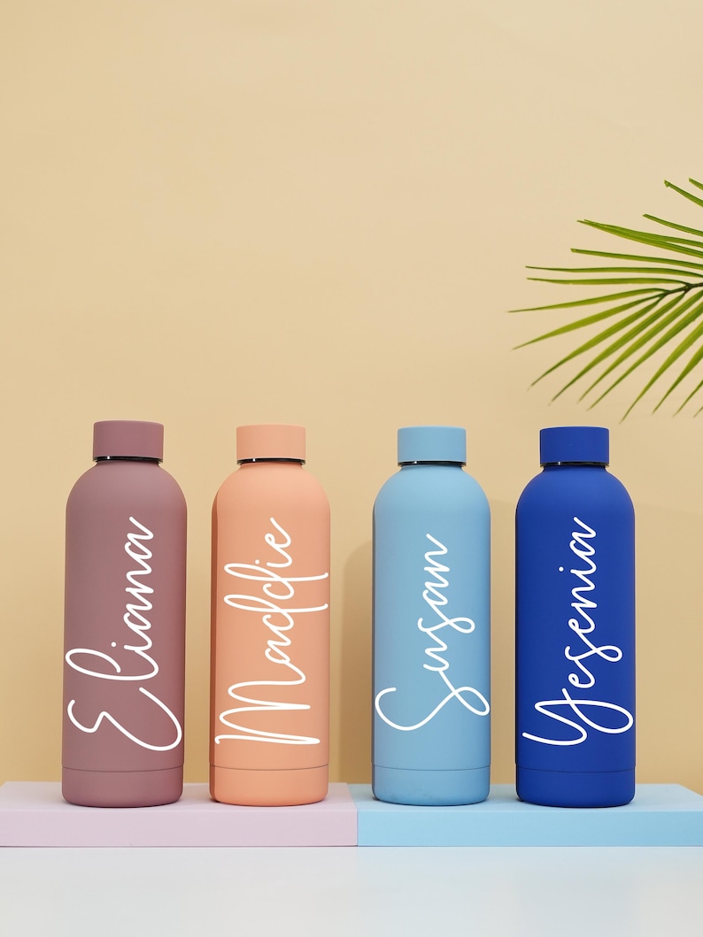 May include: Four personalized water bottles with names "Eliana", "Maclovie", "Susan", and "Yesenia" written in white on the bottles. The bottles are in shades of pink, orange, blue, and dark blue.