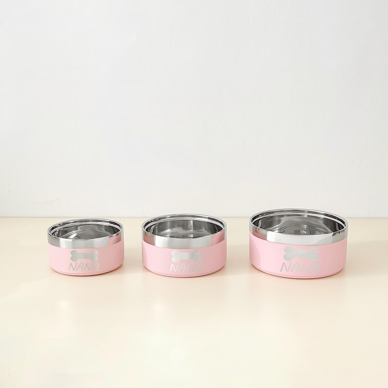 May include: Three pink pet bowls with stainless steel interiors. Each bowl has a silver bone design and the name "NANA" printed on the front. The bowls are in different sizes, suitable for various pet needs.
