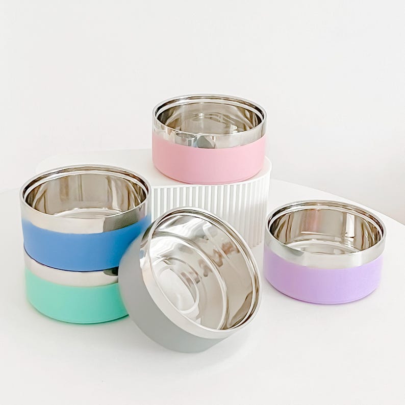 May include: A collection of stainless steel pet bowls with colorful silicone bases. The bowls are in various colors, including pink, blue, green, gray, and purple. The bowls have a shiny, metallic interior and a smooth, matte exterior.