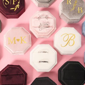 Peut inclure: Diverses boîtes à bagues en velours de différentes couleurs, dont rose, gris et bordeaux. Certaines boîtes présentent des lettres dorées, comme des initiales ou une seule lettre. Plusieurs boîtes contiennent des bagues.