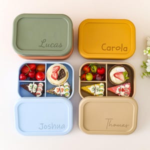 Può includere: Quattro lunch box colorati con coperchi, ognuno con un nome: Lucas, Carola, Joshua e Thomas. Le scatole sono piene di frutta e dessert. Le scatole sono nelle tonalità del verde, giallo, blu e beige.
