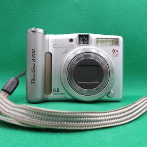 May include: Silver Canon PowerShot A700 digital camera with a 6x optical zoom lens. The camera has a 6.0-megapixel resolution and a silver and gray wrist strap. The camera is on a green background.