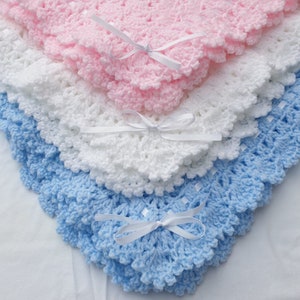 May include: Three crocheted baby blankets stacked on top of each other. The top blanket is pink, the middle blanket is white, and the bottom blanket is light blue. Each blanket has a white satin ribbon tied in a bow at the corner.