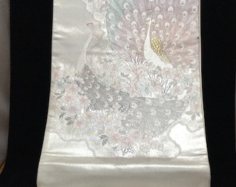 May include: Cream-coloured fabric featuring a detailed peacock design. The design shows two peacocks with white bodies and gold accents, surrounded by floral patterns in shades of pink, grey, and silver. The fabric has a satin-like sheen.