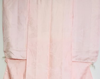 Könnte beinhalten: Ein blasses rosa Seidenkimono mit einem dezenten Blumenmuster. Der Kimono hängt an einem Kleiderbügel und hat einen traditionellen japanischen Stil.