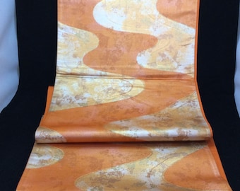 May include: A folded fabric featuring a flowing, wave-like design in orange and gold. The textile has a metallic sheen, with layers of the pattern visible. The colour scheme includes shades of orange, gold, and white, creating an attractive textile.
