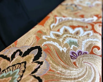 Könnte beinhalten: Nahaufnahme eines dekorativen Stoffes mit einem komplizierten Blumenmuster. Das Design weist Gold-, Orange-, Weiß- und Lilatöne auf. Die Textur und das detaillierte Design des Stoffes deuten darauf hin, dass er für Kleidung oder Heimdekoration verwendet werden könnte.