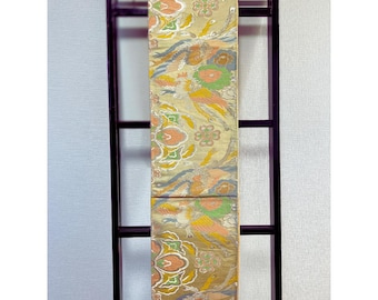 May include: A decorative textile featuring a repeating pattern of colourful birds and floral motifs in shades of yellow, orange, green, and blue. The fabric is displayed vertically on a black wooden frame.