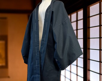 Op de afbeelding: Een donker marineblauwe jas in kimono-stijl met wijde mouwen, tentoongesteld op een mannequin. De jas heeft witte stiksels langs de voorranden en naden. Het kledingstuk is gemaakt van een textuurstof, wat duidt op een traditioneel ontwerp.
