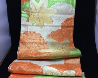May include: A folded textile with a repeating pattern of gold, orange, white, and green leaf and cloud designs. The fabric has a shimmering, textured appearance. The colors are vibrant and the design is intricate.