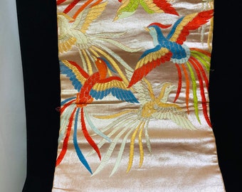 May include: A beige textile with embroidered phoenixes in flight. The birds feature vibrant colours including red, blue, green, and yellow. The fabric is folded, revealing a glimpse of the underside.