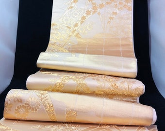 May include: Rolled cream-coloured fabric with a gold floral pattern. The design features stylized flowers and foliage. The fabric has a subtle sheen, possibly silk or satin. The overall aesthetic is elegant and traditional, with a Japanese influence.