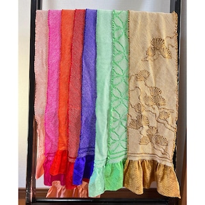 May include: A collection of eight colorful woven scarves with fringed edges. The scarves are arranged in a rainbow pattern, with the colors ranging from peach to gold.
