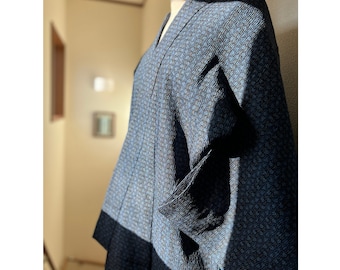 Op de afbeelding: Een blauwe kimono met patroon en een getextureerd oppervlak. De kimono heeft een V-hals en lange mouwen. Het onderste deel van de kimono is effen donkerblauw van kleur. De kimono wordt op een mannequin getoond.