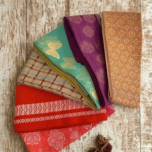 Puede incluir: Una colección de piezas de tela coloridas, incluyendo rojo, turquesa, morado y dorado, dispuestas sobre una superficie de madera clara. Las telas presentan diversos patrones, como diseños florales, de mariposas y geométricos. Los textiles están doblados y apilados.