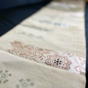 Può includere: Tessuto color crema con un motivo floreale e di nuvole ripetuto. Il tessuto presenta una fascia decorativa con un motivo floreale marrone e un bordo argentato. L'estetica generale è delicata e ornata, suggerendo un tessuto.