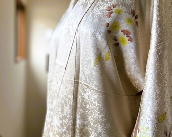 May include: A white silk kimono with a subtle pattern of small white dots. The kimono is decorated with a floral design in shades of yellow, brown, and white.
