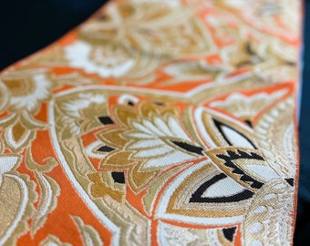 May include: Close-up of a patterned fabric with an orange background and gold, white, and black floral designs. The fabric has a textured appearance and is folded, revealing the intricate details of the pattern.