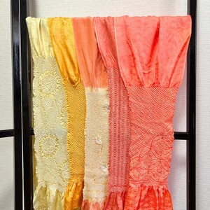 May include: A collection of colorful, patterned fabric panels in shades of yellow, orange, and coral, hanging on a black metal rack. Each panel features unique textures and designs, with ruffled edges at the bottom. The fabrics appear to be lightweight and flowing.