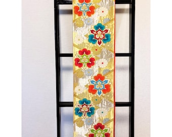 May include: A decorative textile with a gold background and a repeating floral pattern in red, blue, and white. The fabric is displayed vertically against a black frame, showcasing its intricate design and vibrant colours.