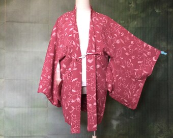 May include: A dusty rose kimono with a floral pattern, displayed on a mannequin. The kimono features a white tie closure and wide sleeves. The floral design is a lighter shade of pink.