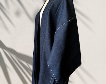 Op de afbeelding: Een donkerblauwe jas in kimono-stijl met wijde mouwen. De jas is aan de voorkant open en heeft witte stiksels langs de naden. Het kledingstuk wordt getoond op een mannequin tegen een neutrale achtergrond, met schaduwen van een plant zichtbaar.
