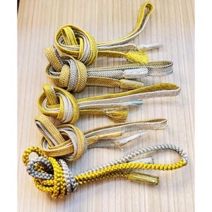 May include: Close-up of several decorative cords with intricate knots. The cords are primarily gold and silver, with tassels at the ends. The cords are arranged on a light-colored wooden surface.