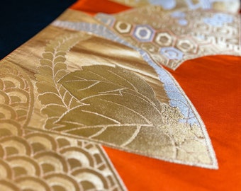 May include: Close-up of a decorative textile with a vibrant orange background. The fabric features intricate gold leaf patterns, including stylized leaves and geometric shapes. The edges are trimmed with a silver-colored border, creating a luxurious appearance.