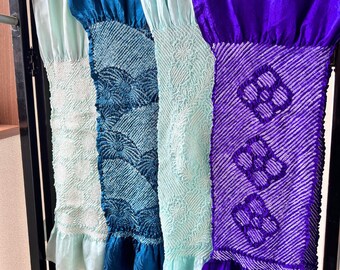 May include: Four colorful, patterned fabric panels hang from a black metal bar. The panels feature shades of teal, blue, and purple, with intricate designs and ruffled edges. The fabrics appear to be silk or a similar material.