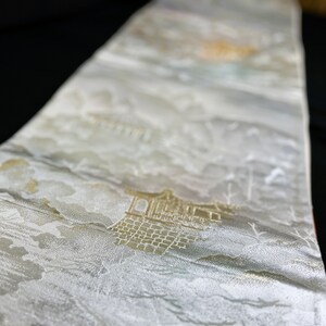 以下が含まれることがあります： 金色の刺繍で日本の建物と山が描かれた白い布。布は上品な光沢があります。