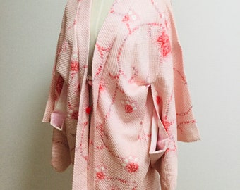 May include: A light pink kimono-style jacket with a textured weave and floral patterns in red and white. The jacket features wide sleeves and pockets, displayed on a mannequin.