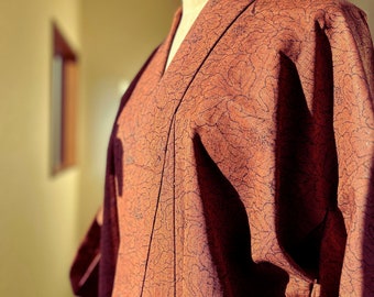 May include: A rust-colored kimono with a floral pattern is displayed on a mannequin. The kimono's fabric has a textured appearance, and the design features dark navy blue outlines. The garment is draped to show the collar and sleeve details.