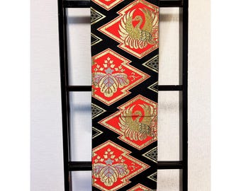 May include: A decorative textile featuring a repeating pattern of red diamond shapes, each with a gold bird or floral design. The background is black, and the edges are trimmed with gold. The textile is displayed vertically.