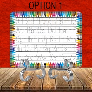 May include: A white learning board with the text "OPTION 1" at the top. The board has the alphabet and numbers 0-9 printed on it. The board is supported by a silver metal stand. The border is decorated with colorful pencils.