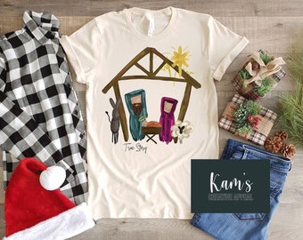 May include: A beige t-shirt with a hand-drawn illustration of the nativity scene. The illustration features a simple wooden shelter, a donkey, a sheep, Mary, Joseph, and a star. The text "True Story" is written below the scene.