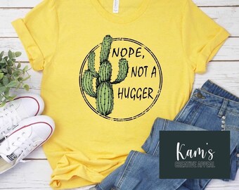 Puede incluir: Camiseta amarilla con un gráfico de cactus verde y el texto "NOPE, NOT A HUGGER".