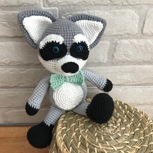 Puede incluir: Un juguete de peluche de mapache tejido a crochet con una pajarita verde. El juguete está sentado en una cesta tejida.