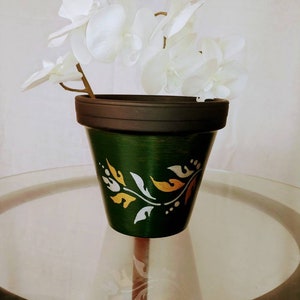 May include: A green ceramic planter with a black rim and gold and silver leaf design. The planter is sitting on a glass table top.