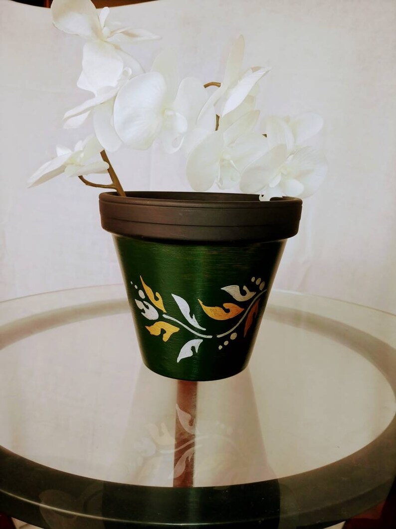 May include: A green ceramic planter with a black rim and gold and silver leaf design. The planter is sitting on a glass table top.