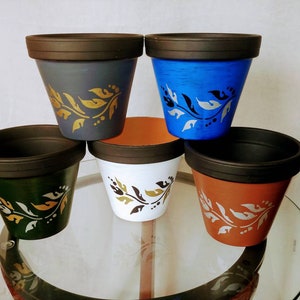 May include: Five terracotta flower pots with black rims. Each pot has a different colour base: grey, blue, white, brown, and green. Each pot has a gold and white floral design.