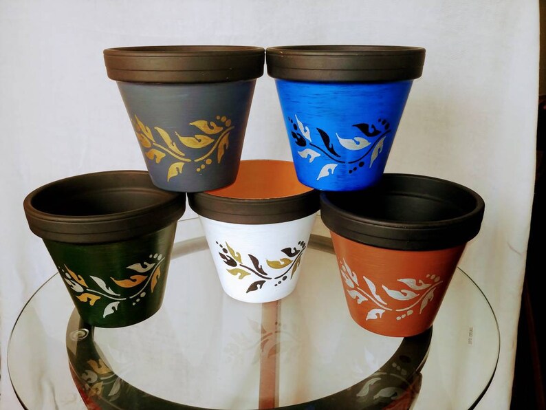 May include: Five terracotta flower pots with black rims. Each pot has a different colour base: grey, blue, white, brown, and green. Each pot has a gold and white floral design.