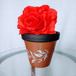May include: A terracotta flower pot with a silver floral design, holding a large red silk rose.