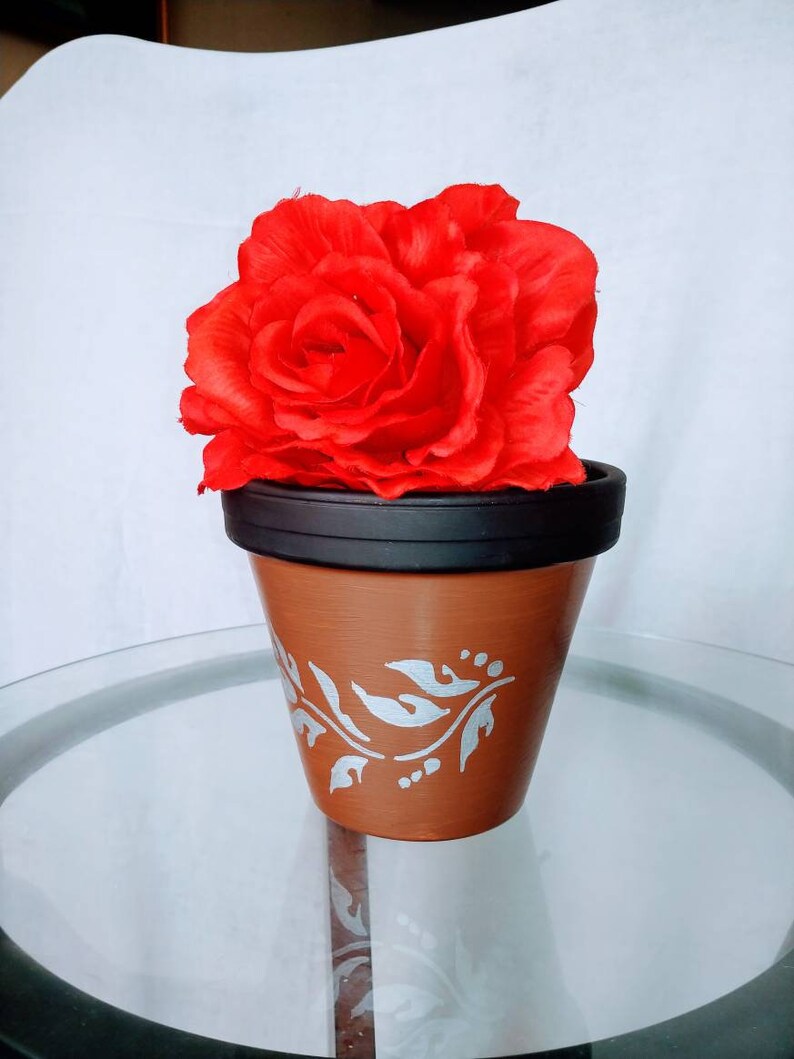 May include: A terracotta flower pot with a silver floral design, holding a large red silk rose.
