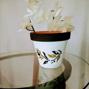May include: White ceramic flower pot with a black rim and gold leaf design. The pot is filled with a white orchid plant.