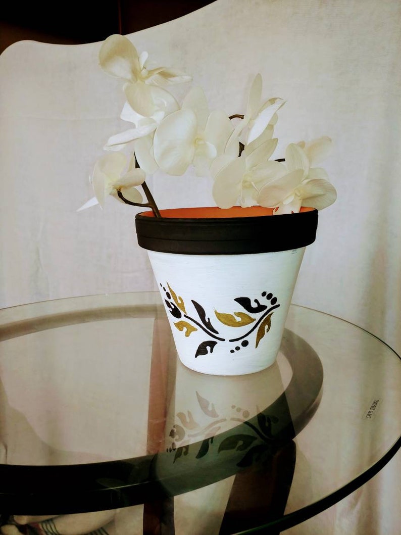 May include: White ceramic flower pot with a black rim and gold leaf design. The pot is filled with a white orchid plant.