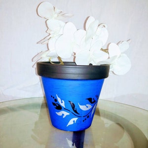 May include: A blue ceramic flower pot with a black rim and a white and black floral design. The pot is filled with a white silk orchid.