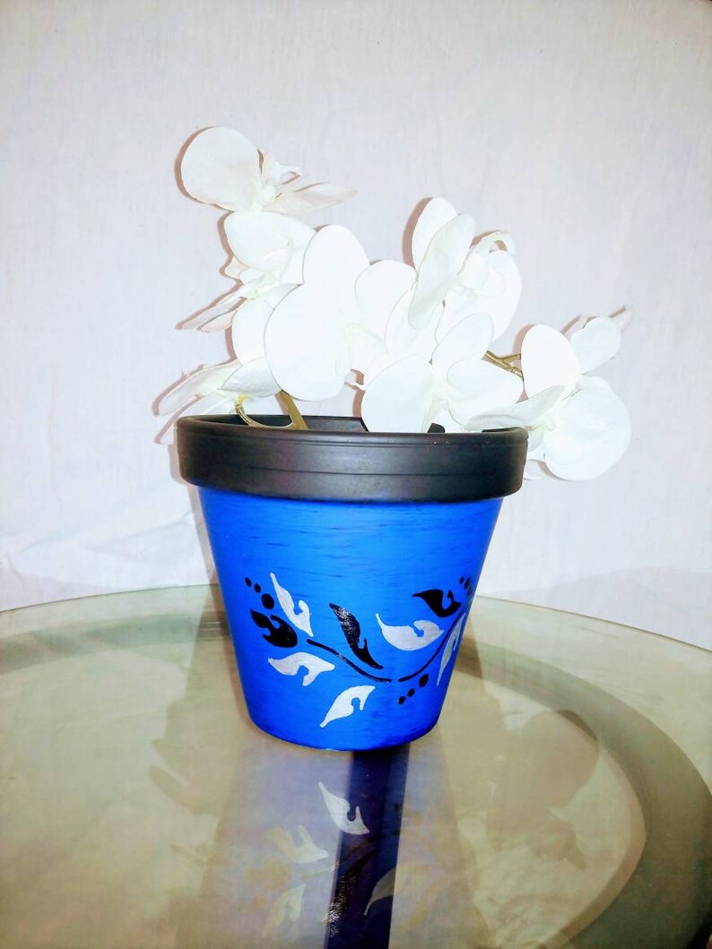May include: A blue ceramic flower pot with a black rim and a white and black floral design. The pot is filled with a white silk orchid.