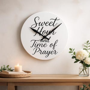 May include: A white, circular wall clock featuring black hands and the script text "Sweet Hour and Time of Prayer". The clock is mounted on a white wall. A candle and a floral arrangement are on a wooden table below.